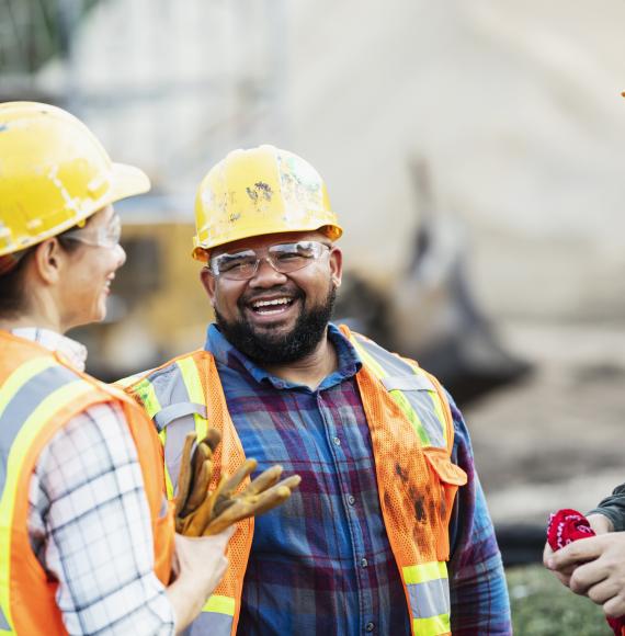 A group of three multi-ethnic workers at a construction site wearing hard hats, safety glasses and reflective clothing, smiling and conversing