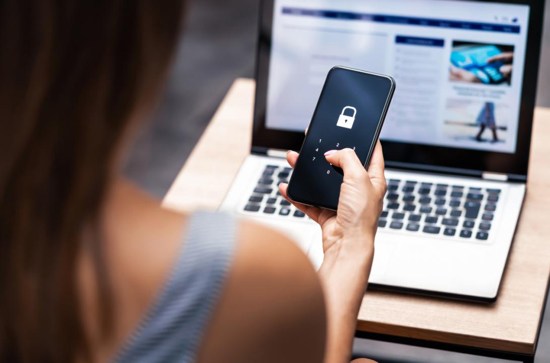 A woman using a laptop and a phone, with the phone showing a padlock on the screen.