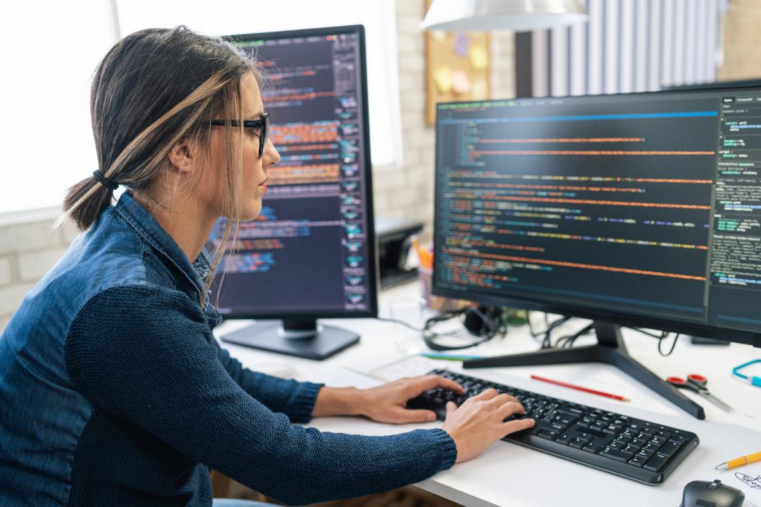 A woman using a computer with two monitors. She is writing code.
