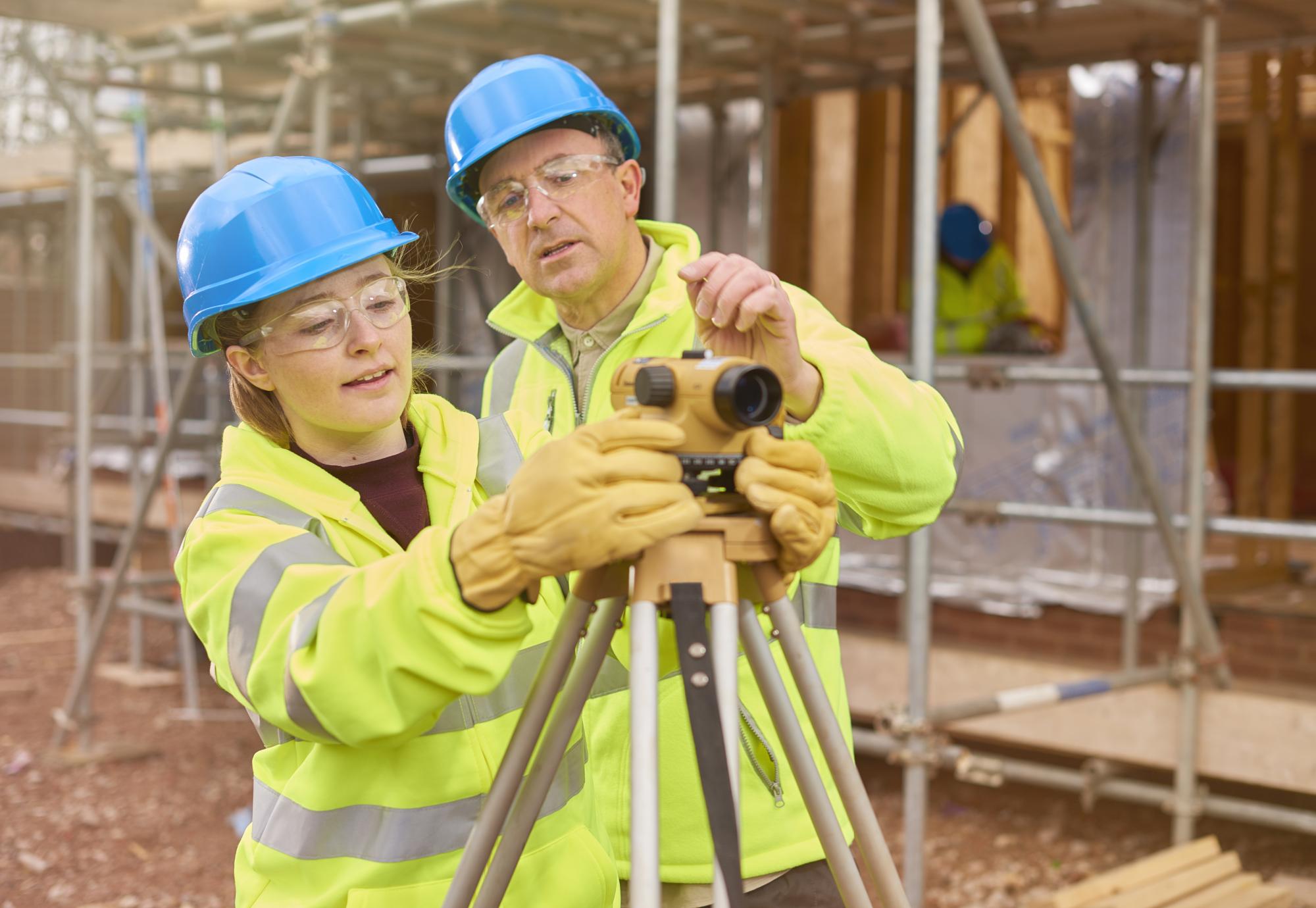 A female construction worker stands behind a builder's level on a building sit