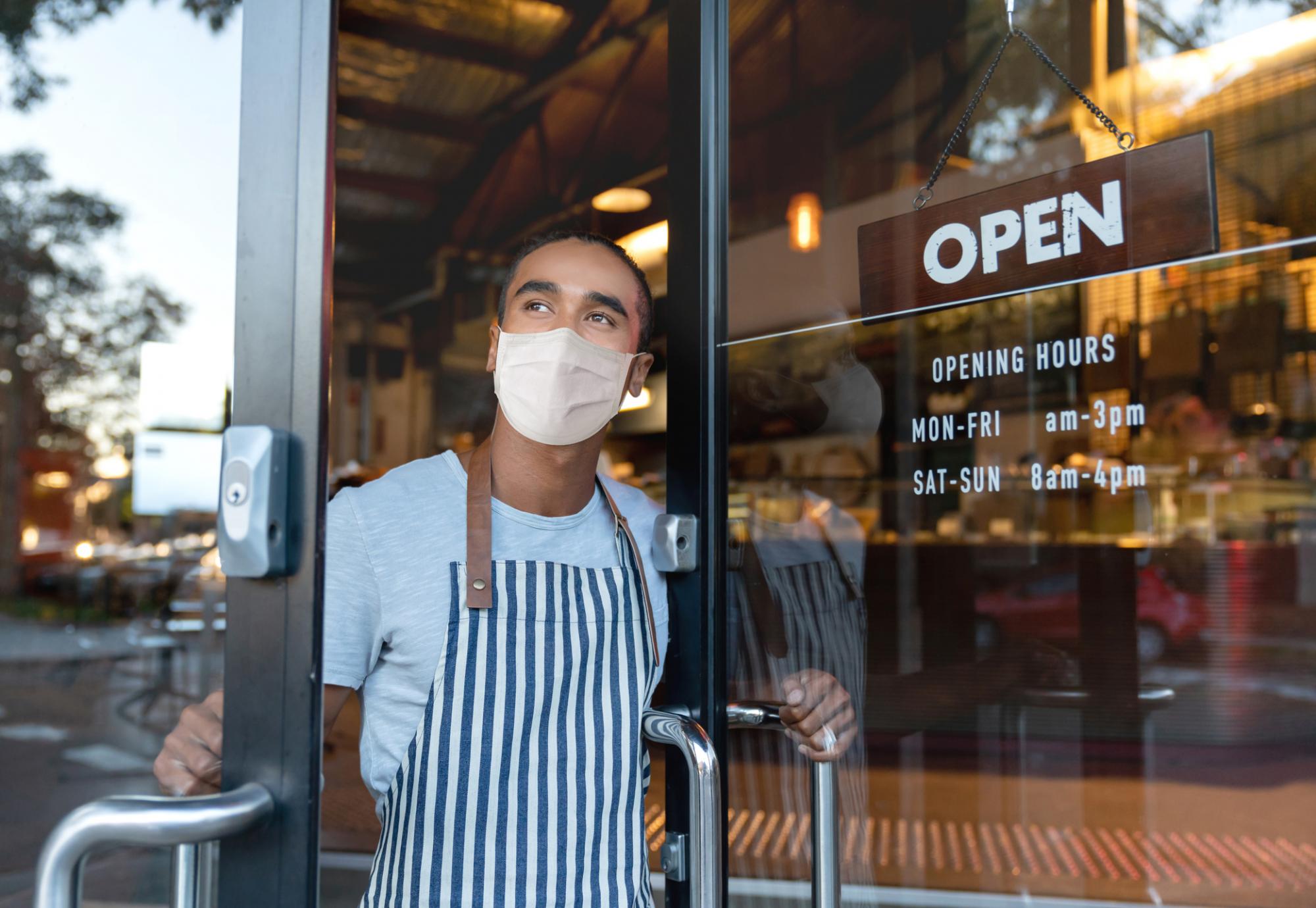 Man in a face covering stands at the door of his business with the door open.