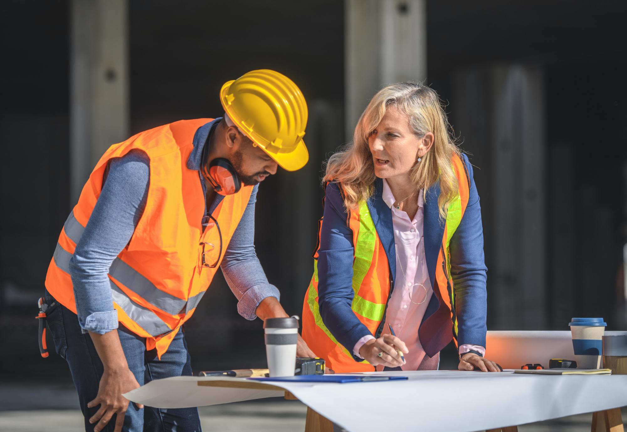 2 construction workers stand over plans of a building.