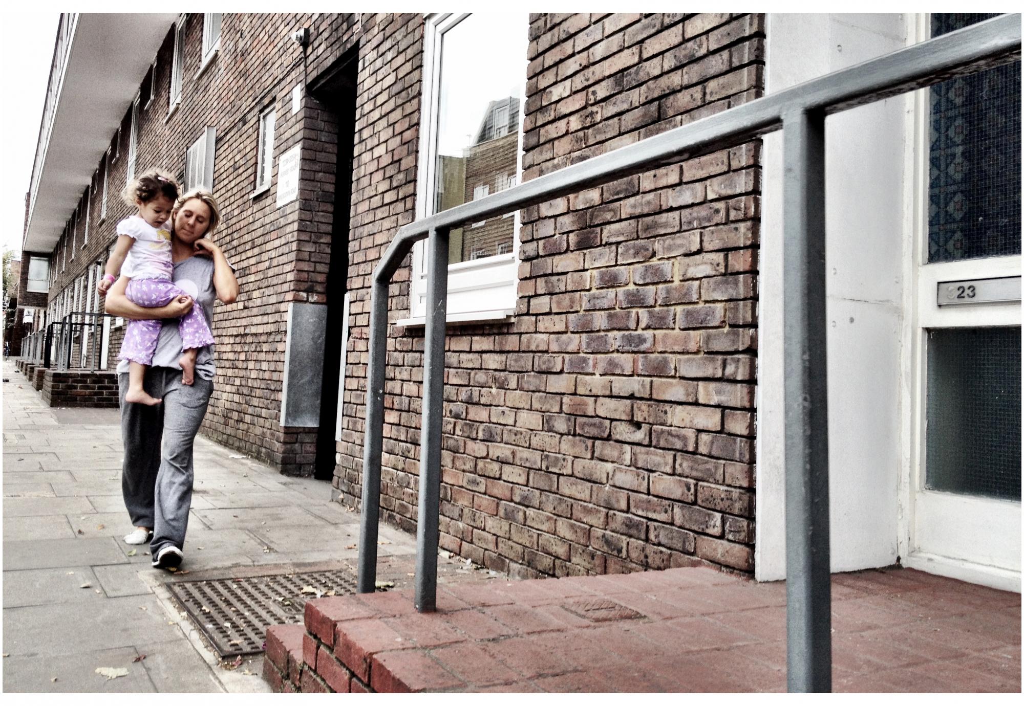 A mother carrying her daughter along the pavement outside a block of council flats 