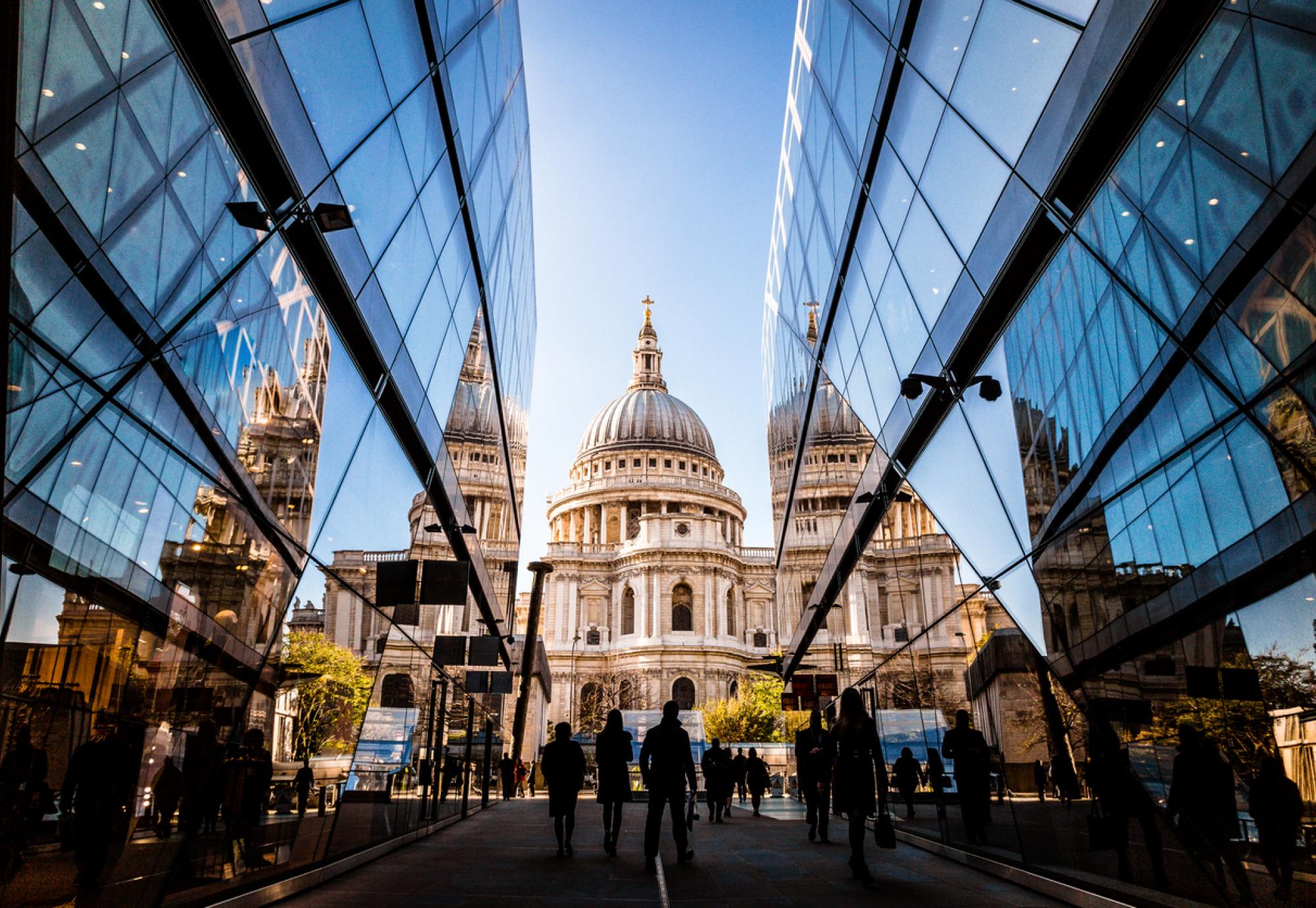 Color image depicting a crowd of people, thrown into silhouette and therefore unrecognisable, walking alongside modern futuristic architecture of glass and steel. In the distance we can see the ancient and iconic dome of St Paul's cathedral