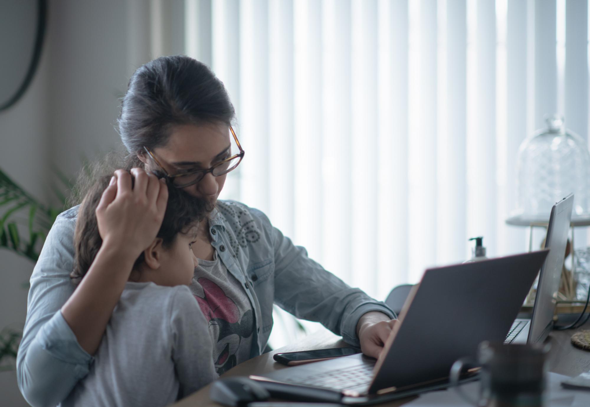 A mother holds her child whilst looking anxious at a laptop