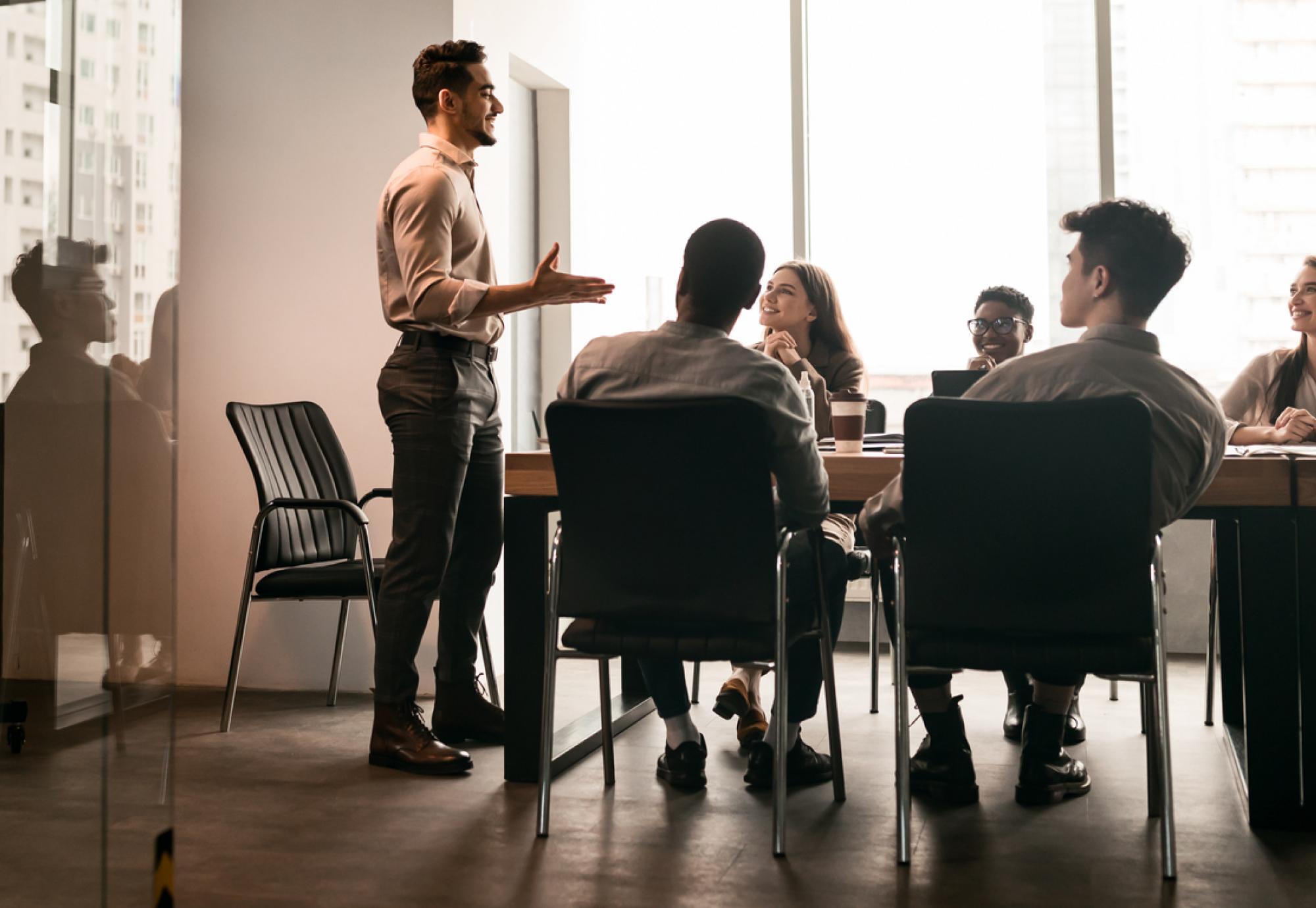 Business Presentation. Smiling Businessman Giving Speech During Seminar With Coworkers In Office, Standing At Desk In Boardroom, Diverse People Sitting At Table And Listening To Speaker