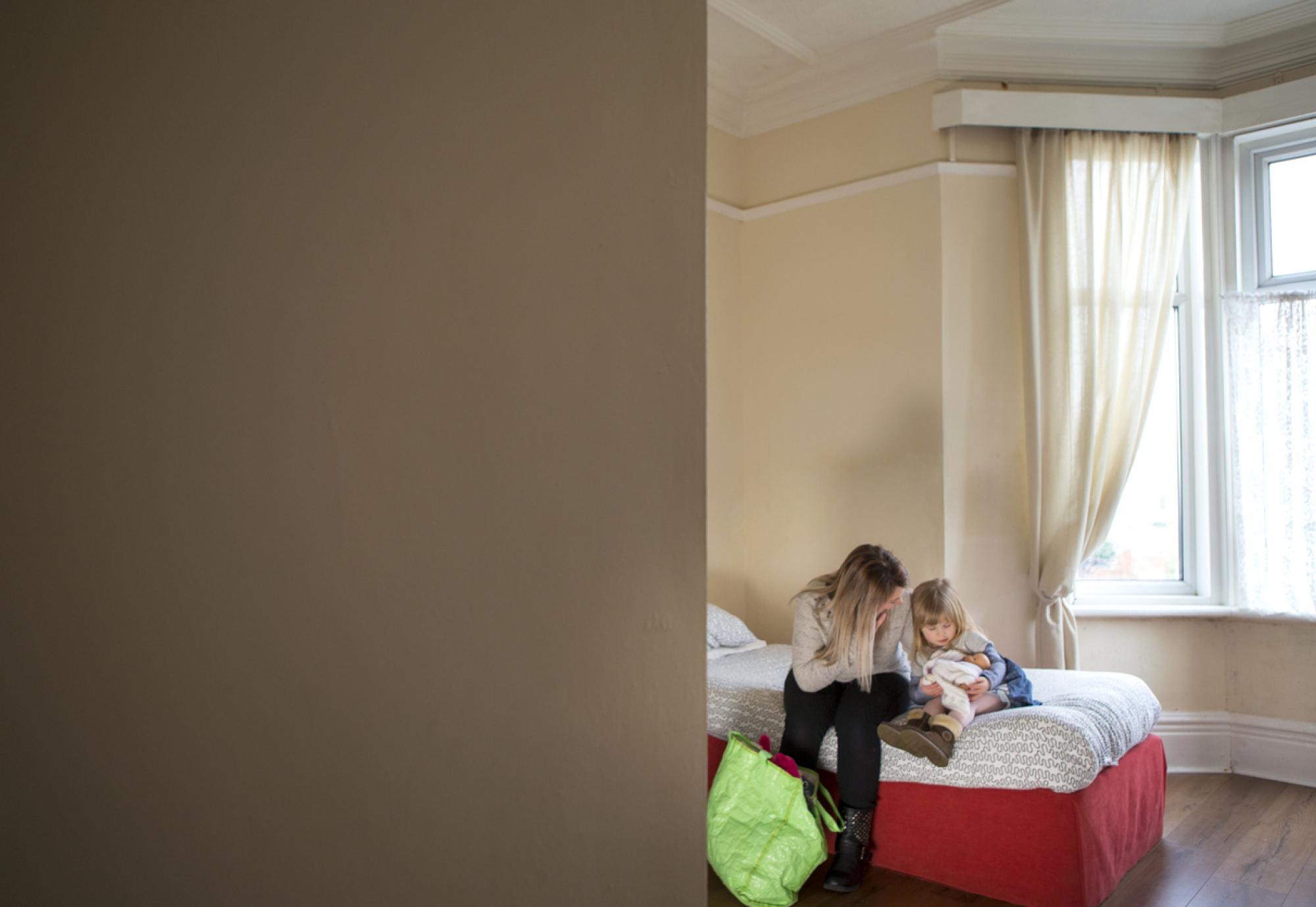 A young mother and daughter sit on a bed in a dingy woman's support centre