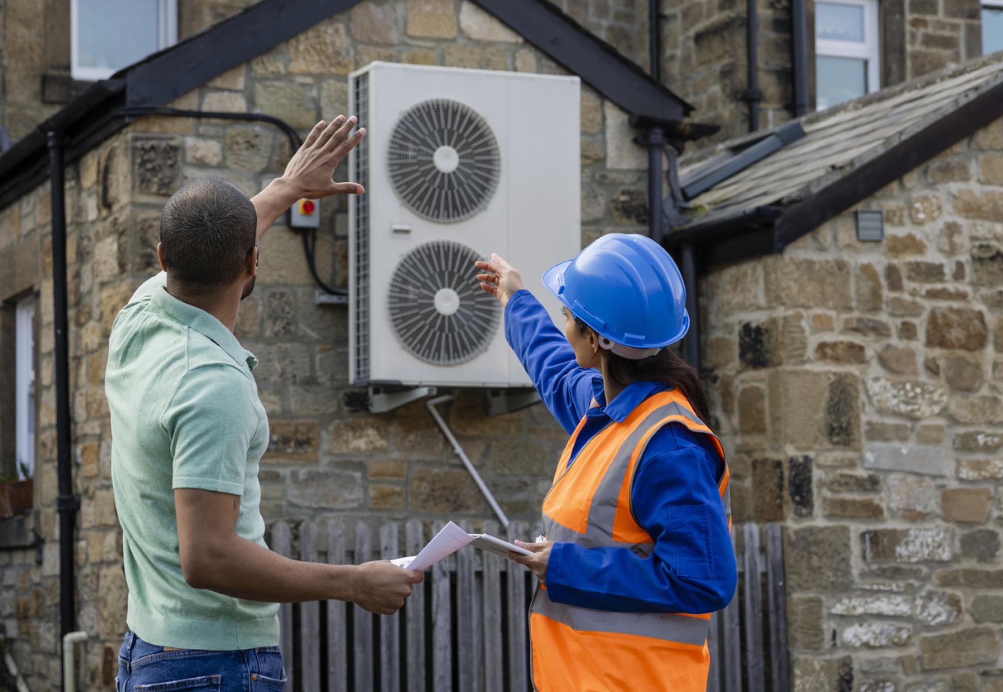 Two people look at a heat source air pump on the exterior of a building