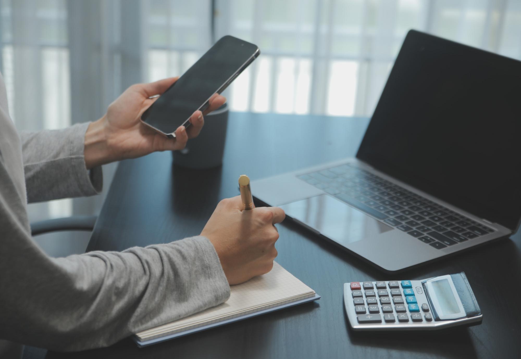 Person working on phone and laptop with a notepad and calculator