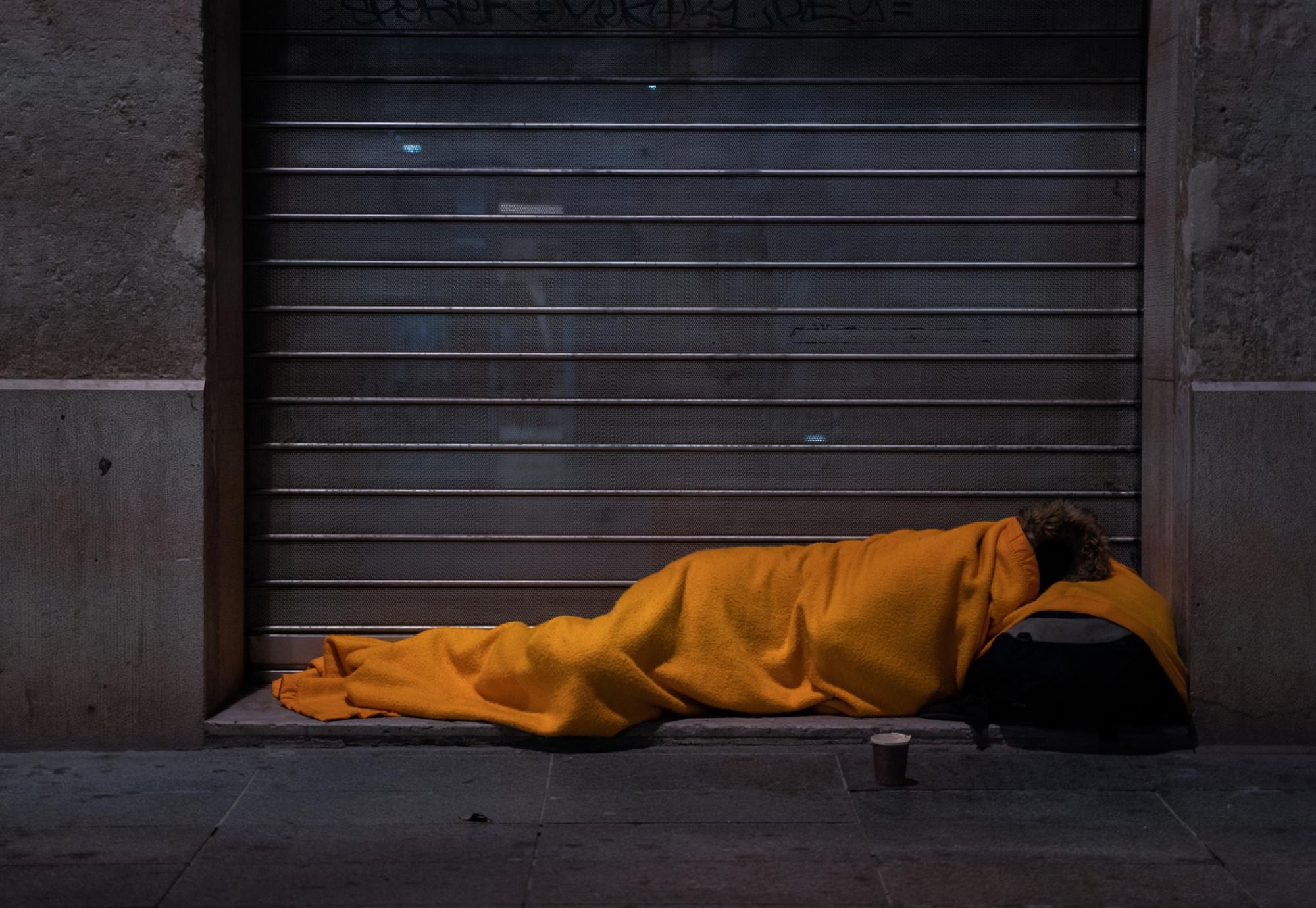 A homeless person sleeps on the street under a blanket