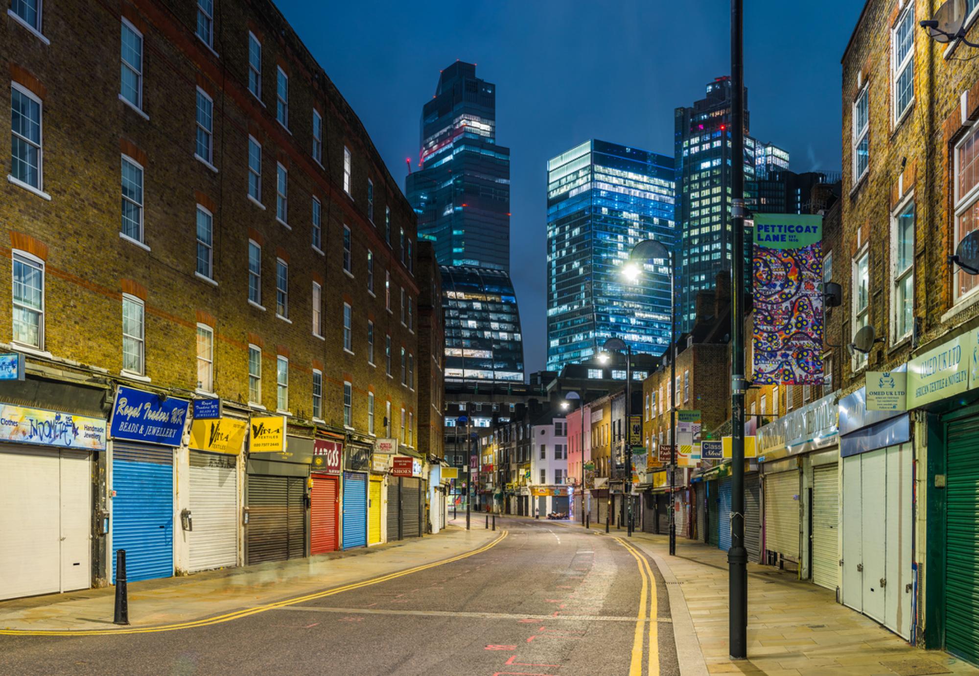 The futuristic towers of the City financial district illuminated at night overlooking the quiet streets and shops of Petticoat Lane Market in Spitafields, London