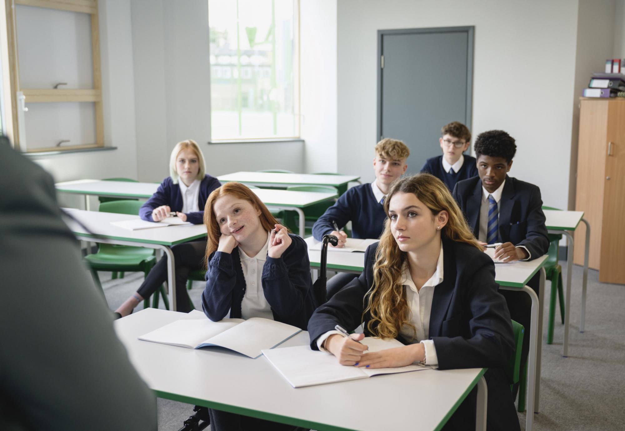 Front view of secondary students wearing uniforms and sitting side by side at desks