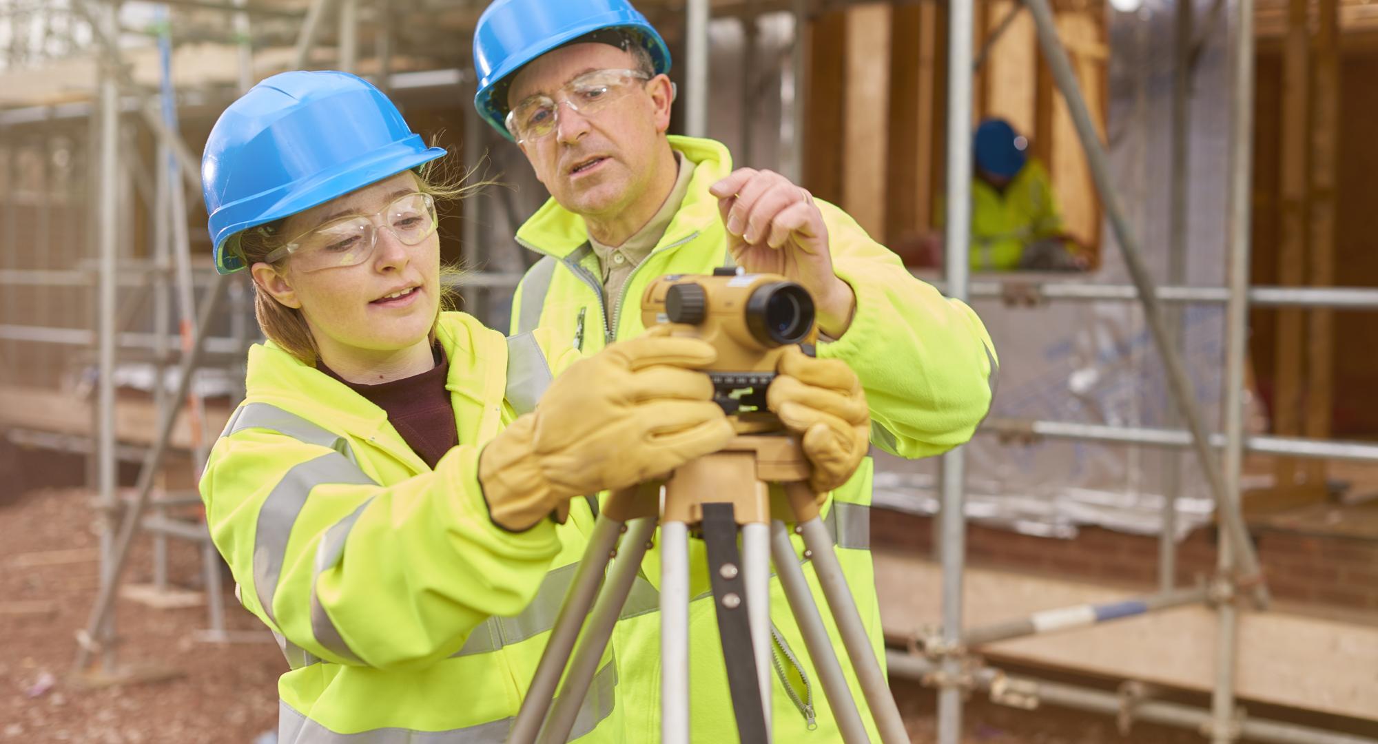 A female construction worker stands behind a builder's level on a building sit