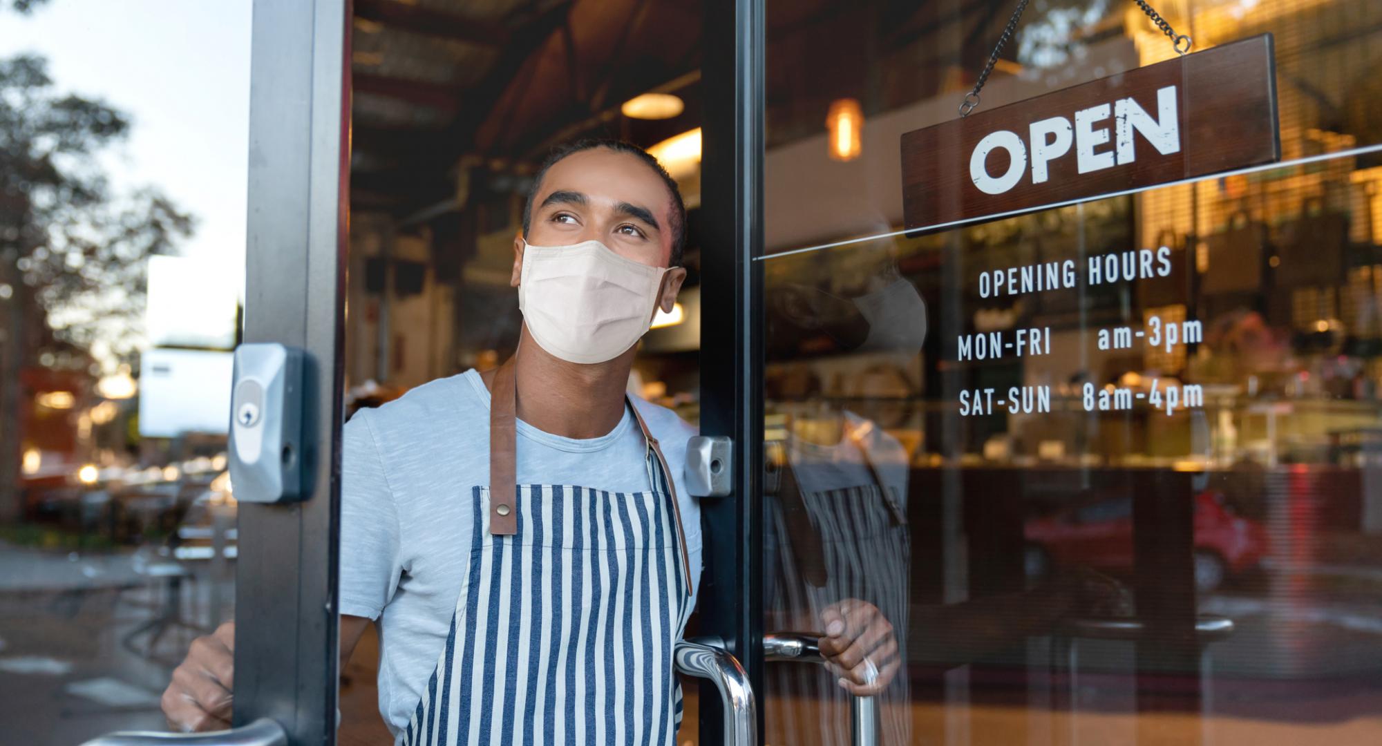 Man in a face covering stands at the door of his business with the door open.