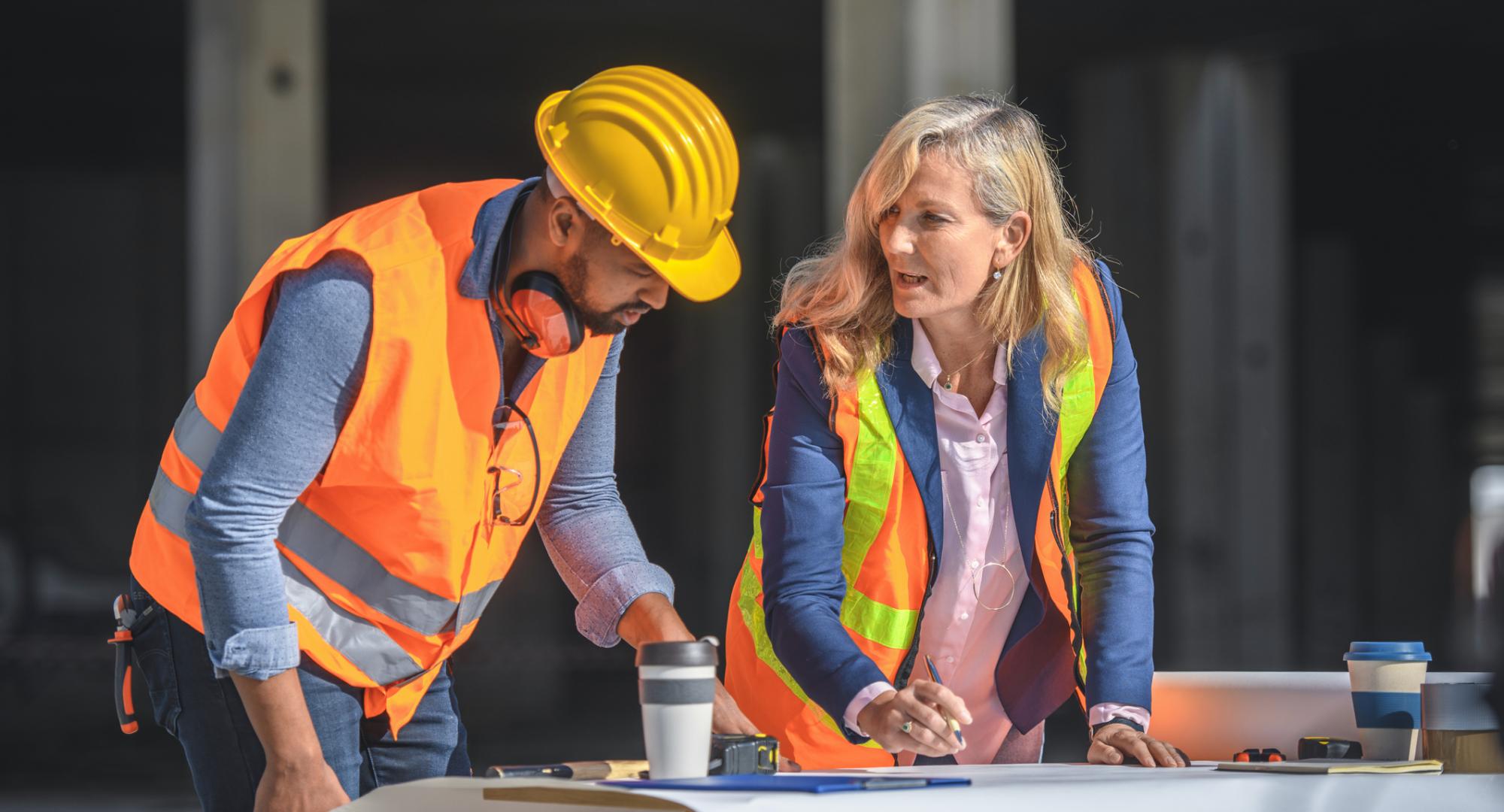 2 construction workers stand over plans of a building.