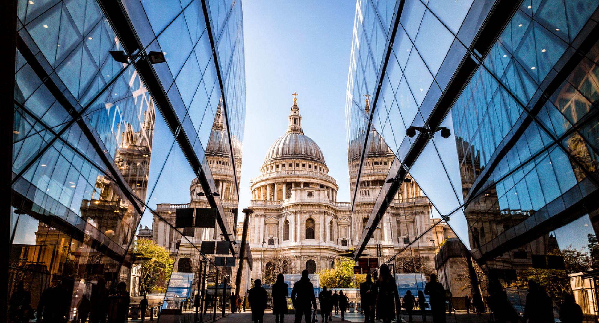 Color image depicting a crowd of people, thrown into silhouette and therefore unrecognisable, walking alongside modern futuristic architecture of glass and steel. In the distance we can see the ancient and iconic dome of St Paul's cathedral