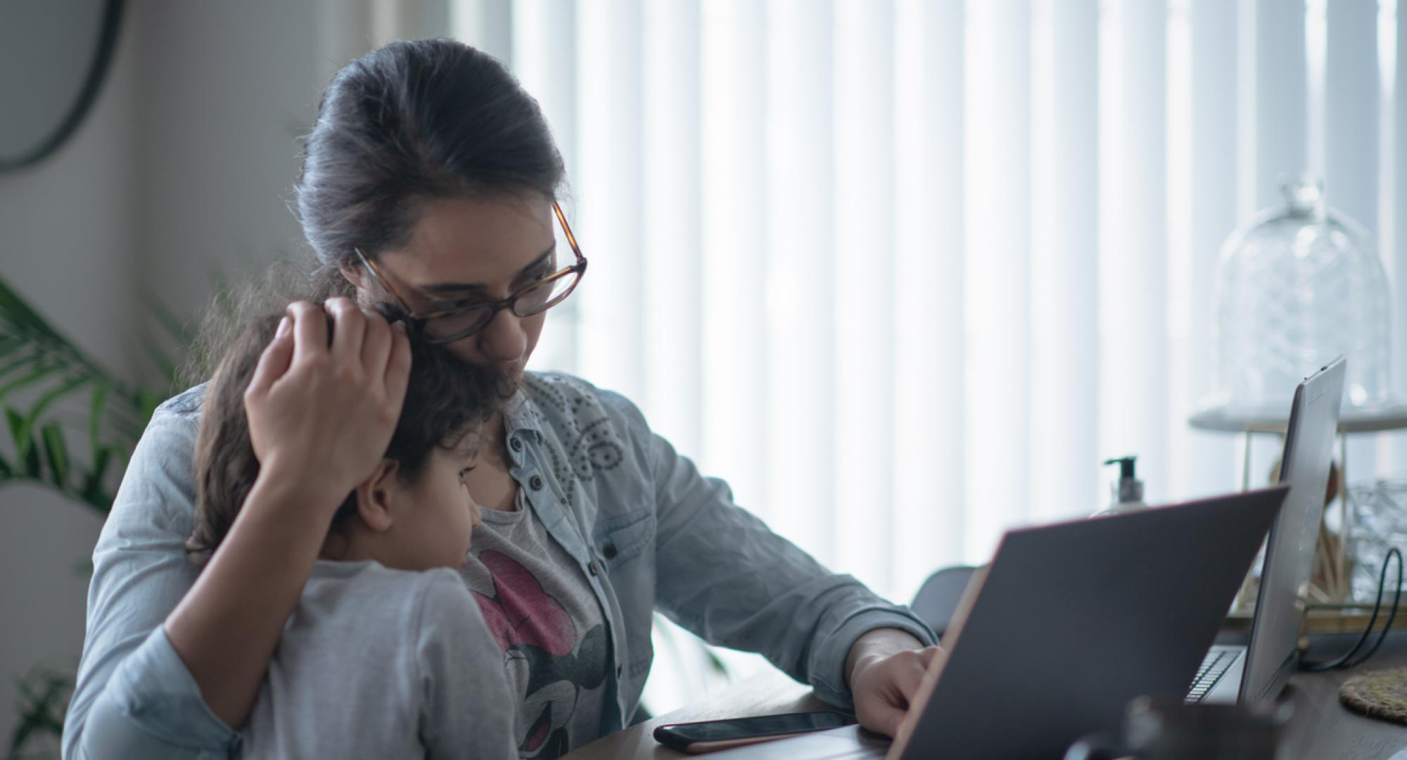 A mother holds her child whilst looking anxious at a laptop