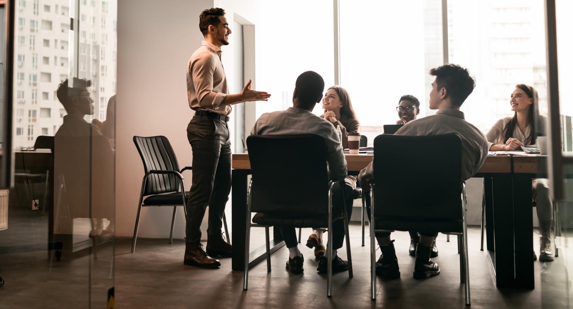 Business Presentation. Smiling Businessman Giving Speech During Seminar With Coworkers In Office, Standing At Desk In Boardroom, Diverse People Sitting At Table And Listening To Speaker