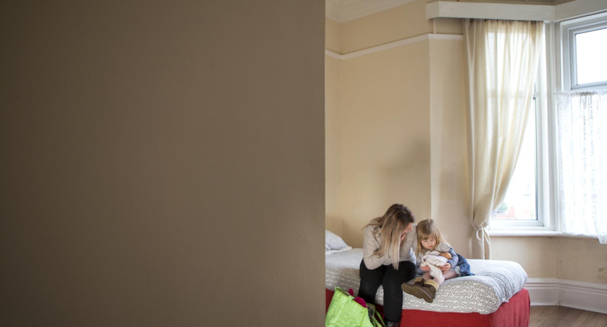 A young mother and daughter sit on a bed in a dingy woman's support centre