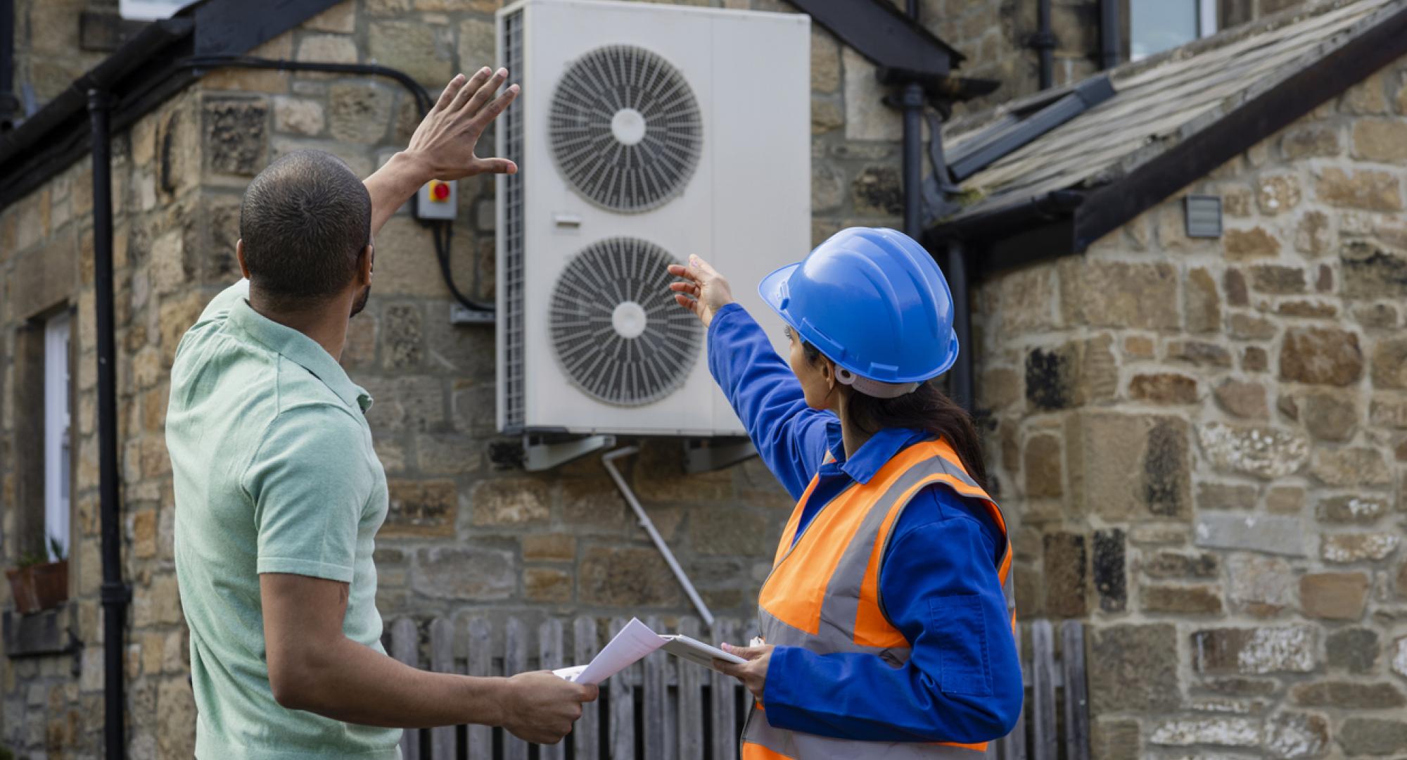 Two people look at a heat source air pump on the exterior of a building