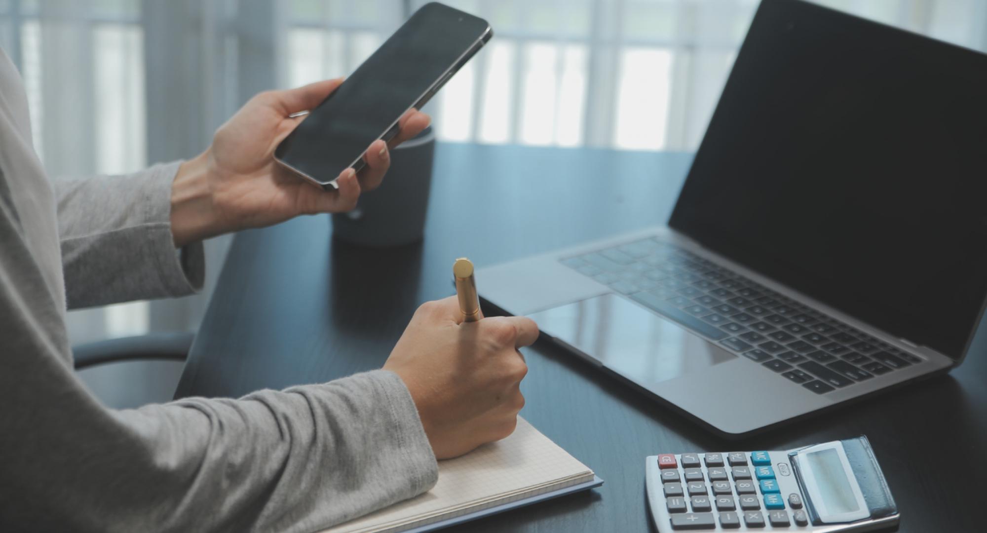 Person working on phone and laptop with a notepad and calculator