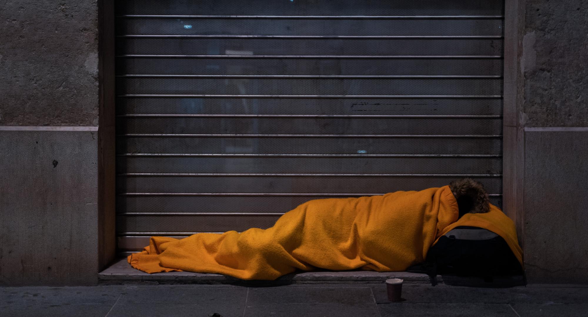 A homeless person sleeps on the street under a blanket