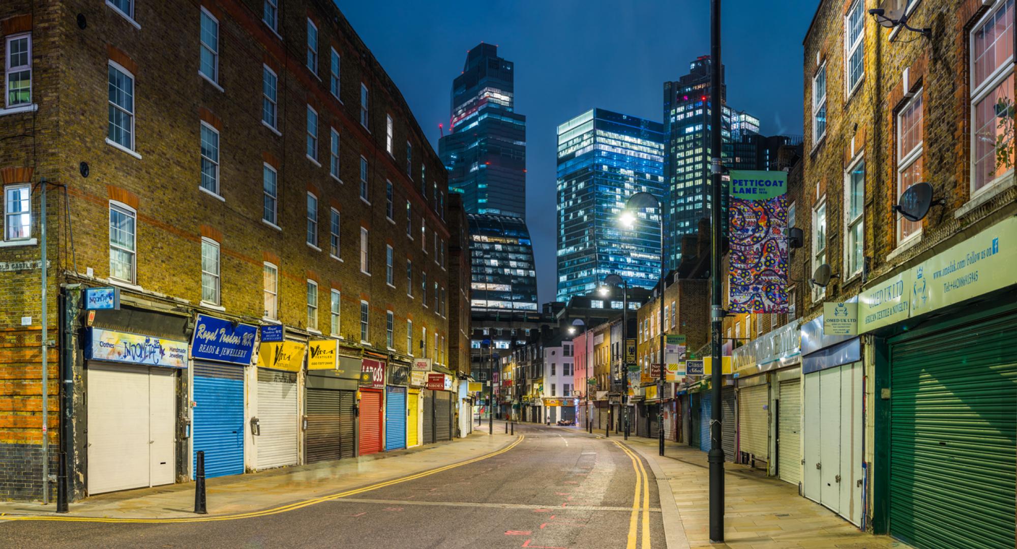 The futuristic towers of the City financial district illuminated at night overlooking the quiet streets and shops of Petticoat Lane Market in Spitafields, London