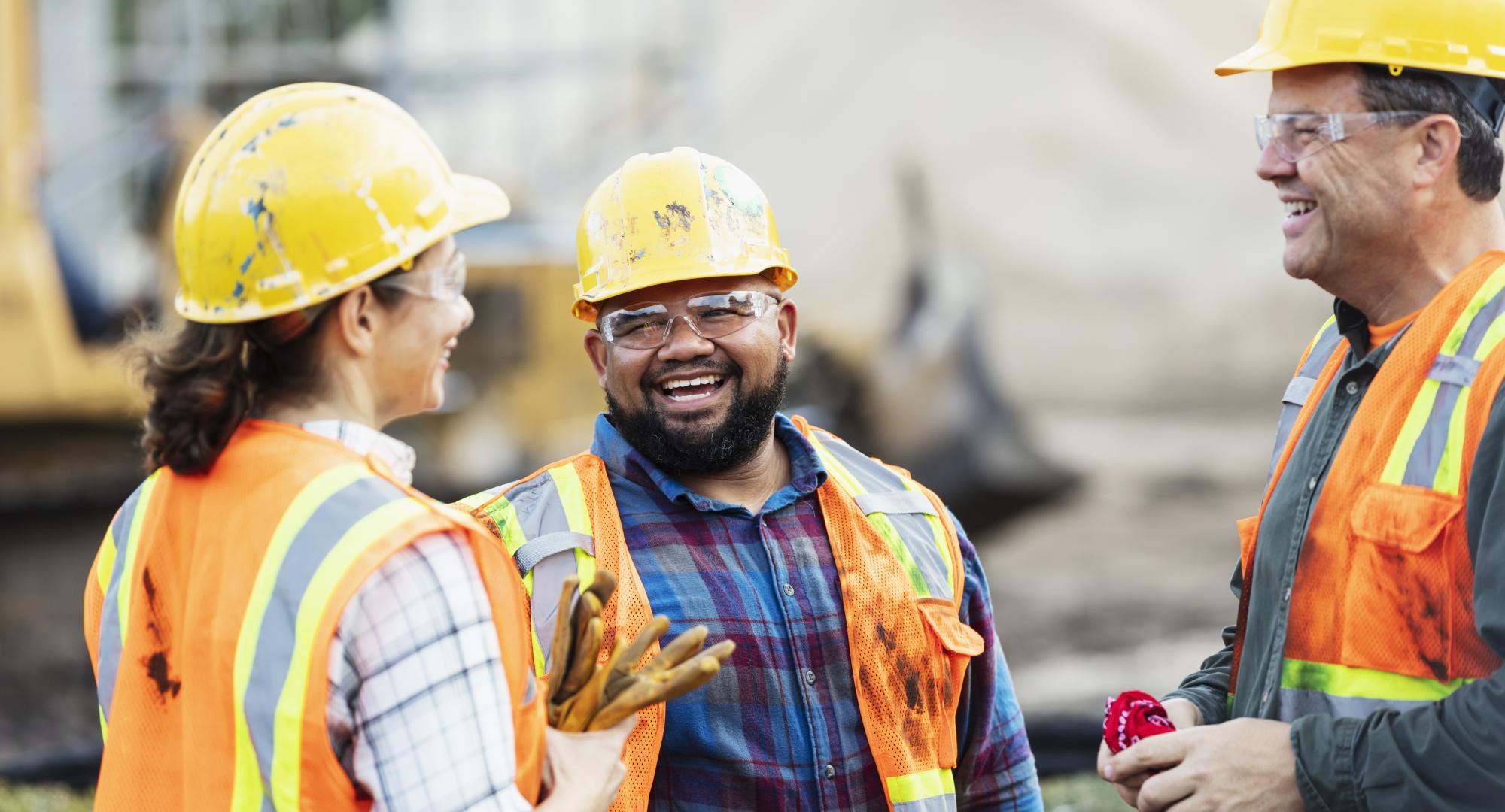 A group of three multi-ethnic workers at a construction site wearing hard hats, safety glasses and reflective clothing, smiling and conversing