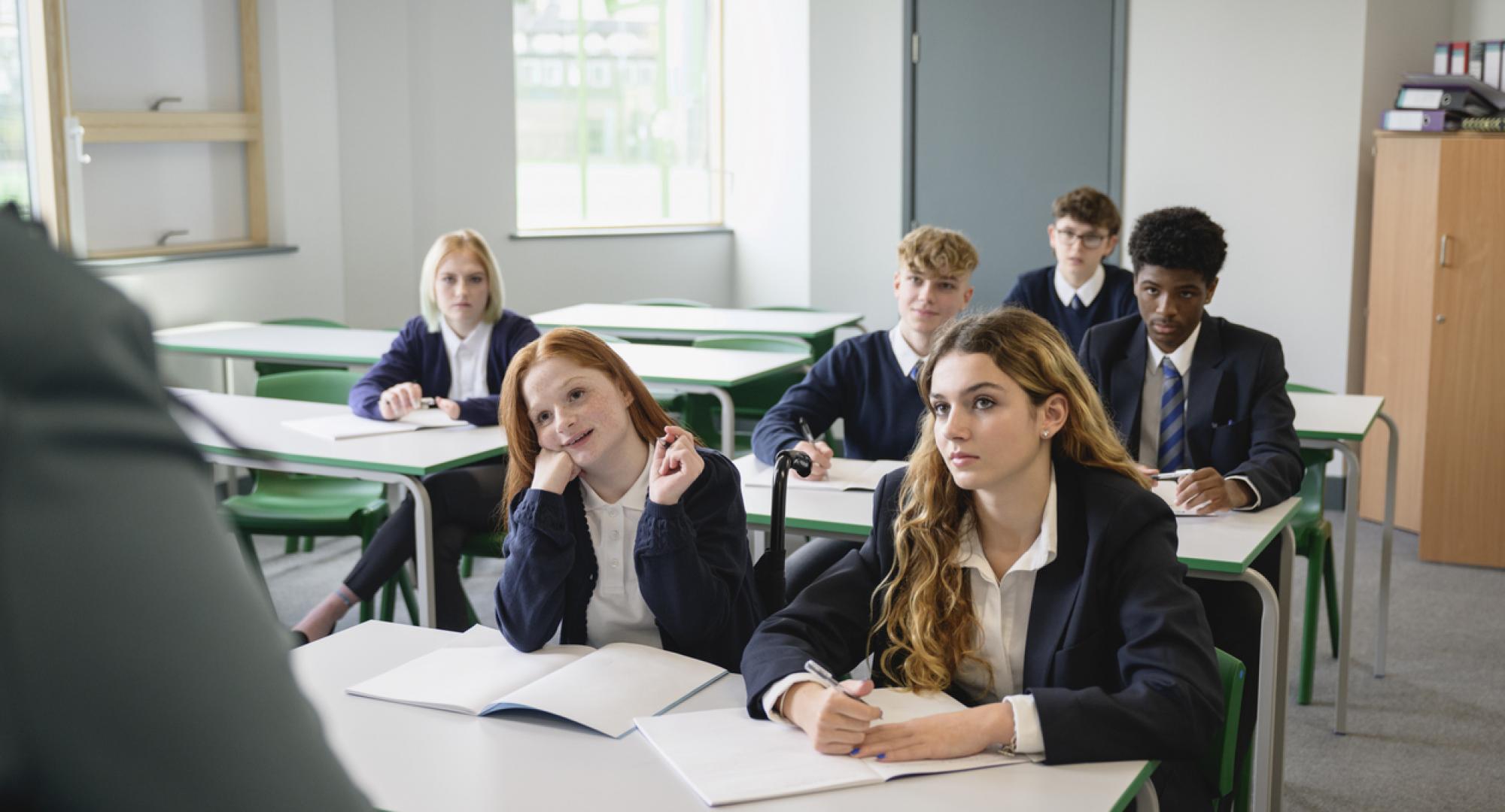 Front view of secondary students wearing uniforms and sitting side by side at desks