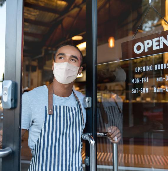 Man in a face covering stands at the door of his business with the door open.