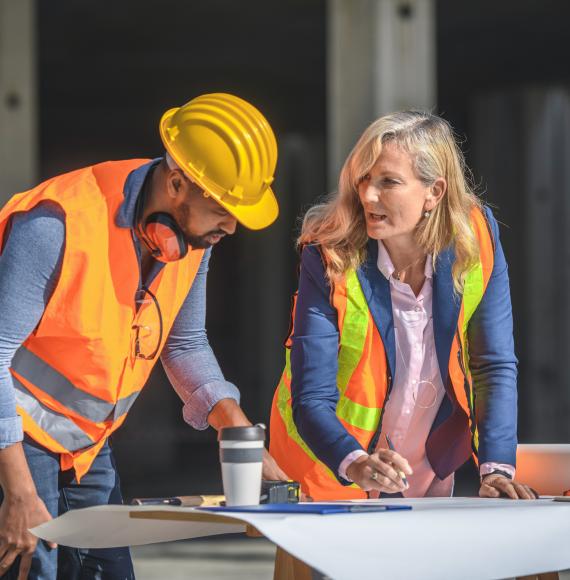 2 construction workers stand over plans of a building.