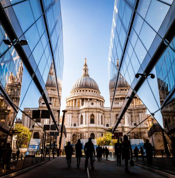 Color image depicting a crowd of people, thrown into silhouette and therefore unrecognisable, walking alongside modern futuristic architecture of glass and steel. In the distance we can see the ancient and iconic dome of St Paul's cathedral