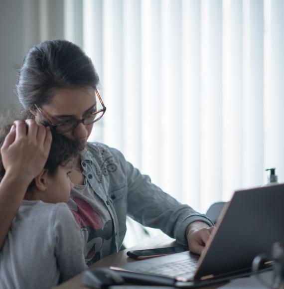 A mother holds her child whilst looking anxious at a laptop