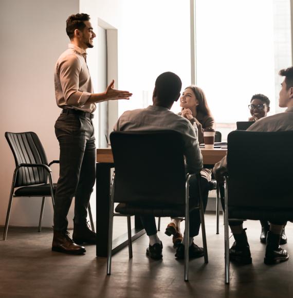 Business Presentation. Smiling Businessman Giving Speech During Seminar With Coworkers In Office, Standing At Desk In Boardroom, Diverse People Sitting At Table And Listening To Speaker