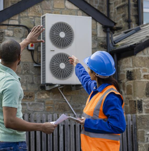 Two people look at a heat source air pump on the exterior of a building