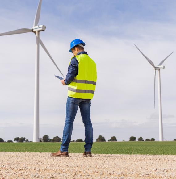 Unrecognizable worker from the renewable energy sector, from the back looking at his digital tablet in a field of wind turbines. Increase in energy prices in the market and alternative energies.