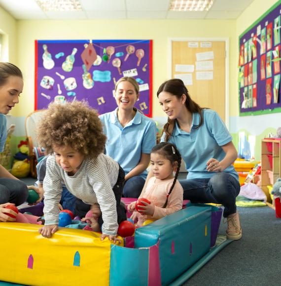 children in a ball pool playing and laughing, behind them sit three nursery teachers